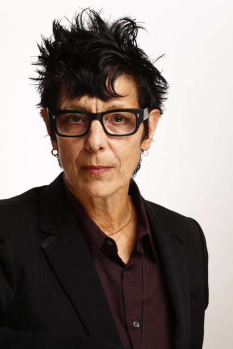 A photo of Elizabeth Streb looking directly into the camera. She has short dark hair and wears dark rectangle glasses. She wears a dark maroon button down top and a black blazer. Photo by Tom Caravaglia.