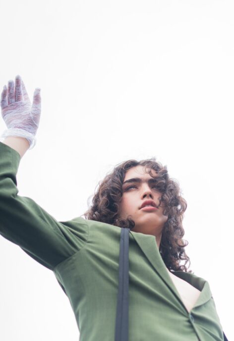 Portrait of Jace in a green blazer against cloudy skies as background. She has curly brown hair and raises her arm towards the sky. She wears a gauzy white glove. Photo courtesy of artist.