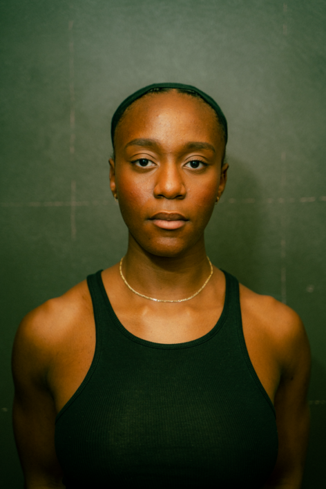 A portrait of woman with mocha skin in front of a green background wearing a black tank top, her hair is tied back in a black head scarf. Photo by Sinematic Studios