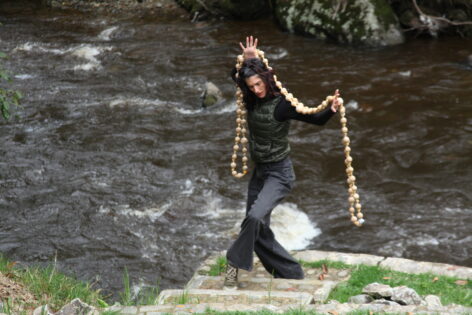 a bio shot of Kay down by the river. She is holding a set of beads behind her and wearing jeans, a long black shirt and green puffy vest. She has dark brown hair, light skin and dark brown eyes. photo by André Daughtry.