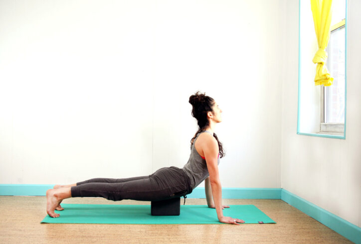 photo for Pilates marketing. Light skin female is in a plank like position with a block supporting her pubic bone for support. Her chest is extended out and up to a window. Kay Ottinger. Photo Courtesy of artist.