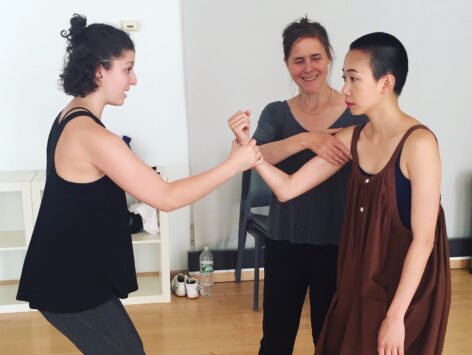 Emily wears a flowly black and white checkered top and a dark skirt. Her right knee is raised higher towards her face. Her left leg is straight on the ground. She appears to be dancing in a living room. Photo courtesy of the artist.