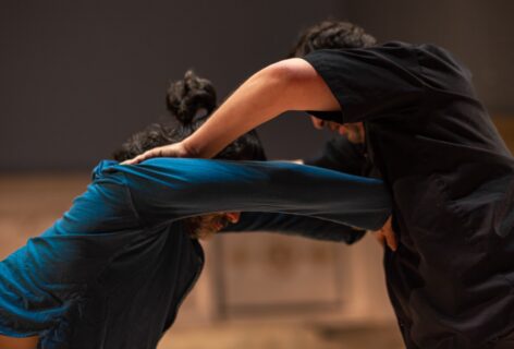 The torsos of two light-skinned males with dark hair and heads bowed are leaning against one another in front of a blurred grey background. One, in a black shirt, is pressing down on the other's shoulders. The other, in a blue shirt, is pressing both hands into the other's chest. Photo by Rachel Keane.