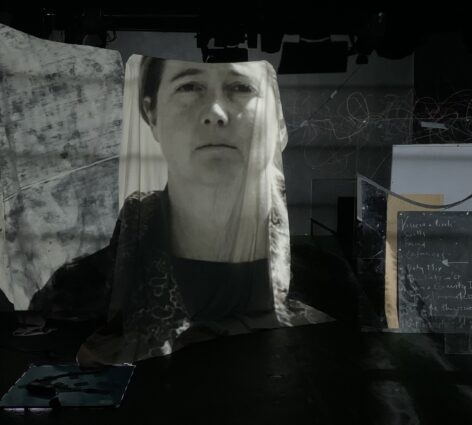 Performance Still of Alys Longley in A Precarious Body of Tilting Maps and Migrant Constellations, University of Auckland, NZ in collaboration with DotDot PopUp Virtual Exhibition. Alys looks straight at the viewer. Their image is projected onto a sheet hanging in a darkened room. Photographer Jeffrey Holdaway. 