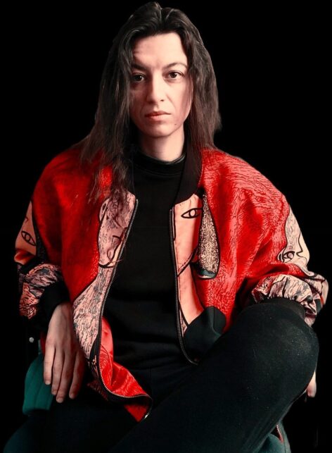 A portrait of Luc Vitk. They look directly into the camera. They have long brown hair and wear a bright red jacket with a pattern of a face down the center split in half by an open zipper. They wear a black top and sit with their legs crossed. Photo courtesy of the artist.