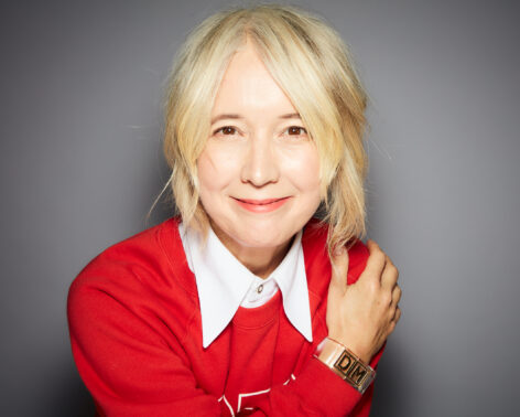 Portrait of Justine Simons smiling warmly at the camera. She has blonde hair and light skin, and wears a red top with a white collar. Photo by Rankin.