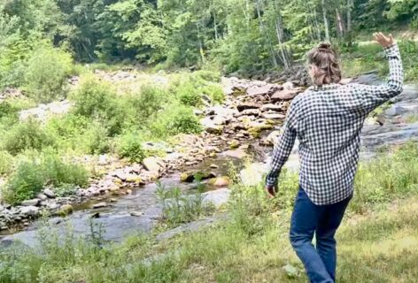 Julie Mayo in nature. Green grass and trees surrounding her as she walks towards a creek. Photo by Mark Richardson.