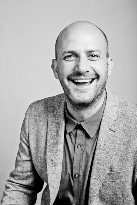 A headshot of Vallejo Gantner. He looks into the camera with a wide smile. He wears a button down top and a blazer. Photo courtesy of the artist.