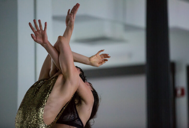 Two light skinned dancers' upper bodies one in front of the other with arms stretched upwards intertwined as if they were vines. Front dancer is wearing a gold sequined leotard opened on the sides and back dancer is wearing a lace navy blue bra. Their faces are not seen but we can tell from seeing part of their heads and hair that they are both tilting back. There is tension in their arms as if they had gotten their arms tied up with one another. Photo by Scott Shaw.