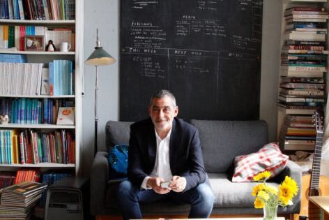 Photo of Lucien Zayan sitting on a couch and smiling at the camera. There is a chalkboard on the wall behind him and bookshelves on either side. Photo by Simon Courchel