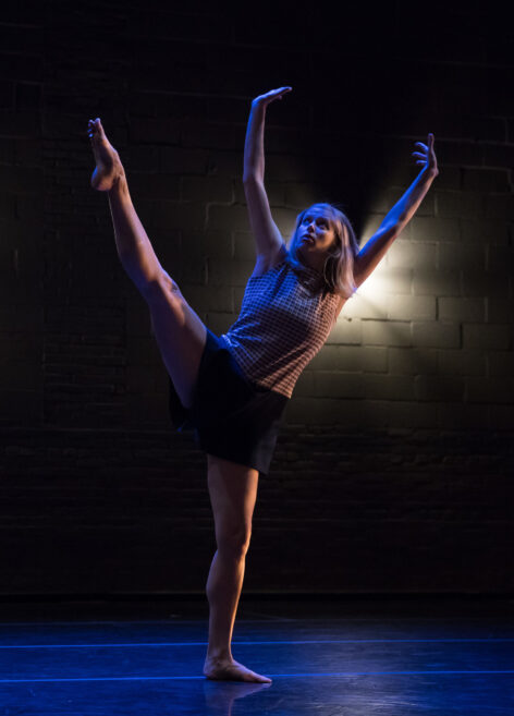 A woman on a dimly lit stage dances with her arms overhead and leg outstretched to the side. Photo by Jonathan Hsu.