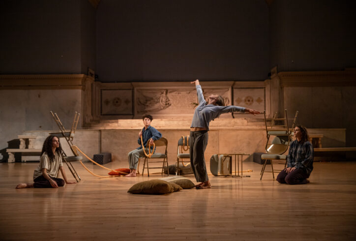 Rebecca Pappas performs for Movement Research at the Judson Church. Four dancers perform using chairs, cushions and rope as props. One dancer in the center stands and arches backwards extending their arms. Three other dancers sit looking at them. Photo by Rachel Keane.