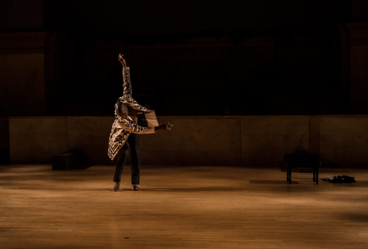 Paul Hamilton. Movement Research at the Judson Church. A dancer wearing a white jacket painted with black abstract patterning reaches one arm straight up, the other to the side. Their gaze is straight down at the floor. Photo by Rachel Keane.