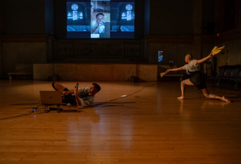 Marie Al Evangelista's work for Movement Research at the Judson Church. A collage of live footage displayed on the projector. One dancer on their side laying on the floor and looking into a camera. Another dancer in a deep lunge holding objects in their outstretched hands. Photo by Rachel Keane.