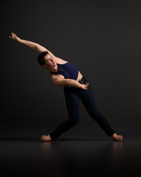 In this dynamic photo, Isabella, barefoot and dressed in a dark sleeveless top and matching pants, leans deeply into a side stretch with one arm extended above their head and the other stretched downward, toward the opposing angle. Her short brown hair and focused expression highlight her strength, balance, and the disciplined yet playful nature of her artistic practice.