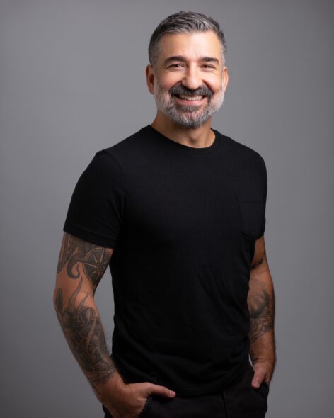 Fabio Tavares standing with his hands in his pockets wearing a black t-shirt and smiling at the camera. Photo by Mike Rubio.