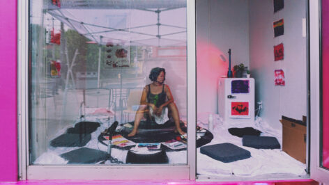 Through a floor-to-ceiling glass storefront, we see an Asian woman with wavy chin-length dark hair sitting bare-legged in a room strewn with seat cushions, a microphone, and an assortment of mysterious, colourful prints and objects. A few prints hang on the wall to her right, as well as an anatomical poster behind her. She sits up with her knees bent, feet planted on the ground, legs spread apart with a crumpled white sheet partially draped over her thighs. She is looking away from the camera. Photo courtesy of the artist.