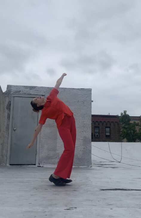 Lucienne wears an all red outfit, as she throws her head up to the sky and arms out. She is on a metallic gray rooftop with the cloudy sky in the background.