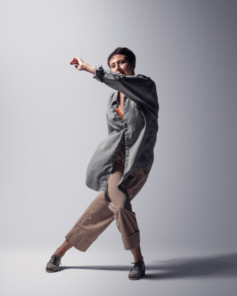 Kaleena wears a gray flowing shirt, khaki pants and gray tap shoes. She is mid-lunge, facing the camera, legs crossing and left arm swinging across body into the air.