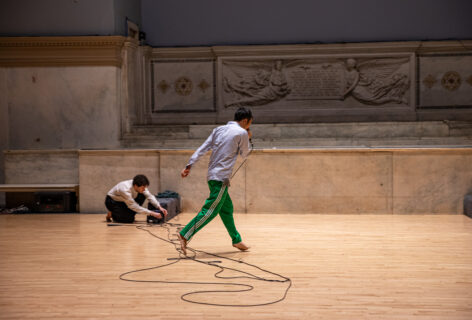 Kensaku Shinohara's performance for Movement Research at the Judson Church. One dancer walks while speaking into a mic with a long chord. Another performer in the background squats down adjusting wires. Photo by Rachel Keane.