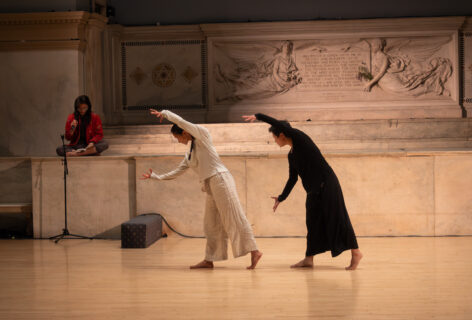 Annie Ming-Hao Wang performs for Movement Research at the Judson Church. Two dancers stand in front of the altar. They are mid step with their torsos curved forward and their arms open and reaching forward. Another performer sits talking into a mic at the altar. Photo by Iki Nakagawa