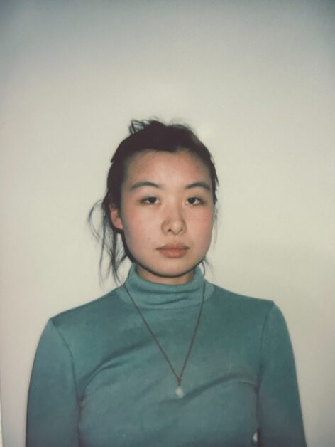 An Asian nonbinary person is standing and smiling. They are wearing green eyeshadow and blush, and their black hair is loosely tied up in a bun. They are dressed in a green turtleneck sweater and a red yarn necklace with a jade pendant. The background is a blank white wall.
