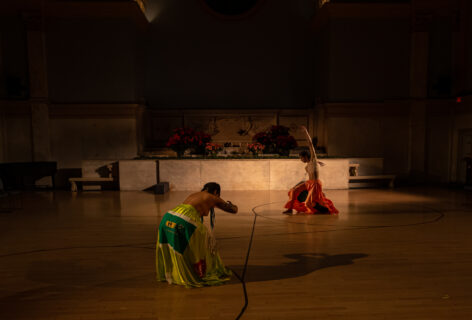 Two dancers moving during Christopher Unpezverde Nuñez Choreography for Movement Research at the Judson Church. Marielys Burgos Meléndez arching forward with her hands out in front of her. She wears a long green skirt. A second dancer lunges forward with one arm reaching upwards. They wear an orange skirt. Photo by Rachel Keane.