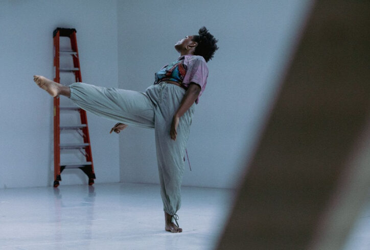 Dark skinned Black woman with a 4c puff of hair looks swings her leg through the air while leaning back. She is wearing a purple graffitied shirt and green pants. Photo by Elyse Mertz