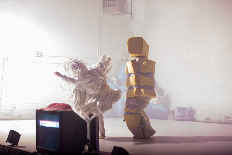 A moment from the performance Face Eaters depicting two figures: one, clad in white mop-like tassels, and an other, wrapped in foam secured by silver duct tape. Photo credit to Elyse Mertz.