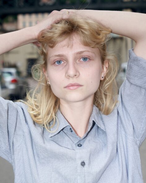 A headshot of Justine Florence with her hands over her head. Her blond hair is partially tied back. She wears a blue plaid button-down and looks at the camera with a neutral expression.