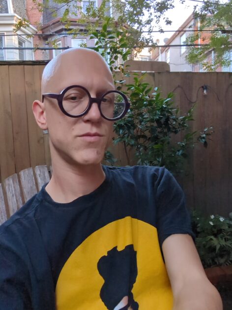 A hairless nonbinary person with large round purple glasses sits in a garden, staring at the camera. Photo courtesy of the artist. 