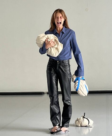 A person stands barefoot on a bag, wearing black leather pants and a blue shirt, screaming with mouth wide open and eyes closed. Holding two fabric- wrapped bundles tightly, one in each arm, while a smaller bundle also lies on the floor. The background is plain and minimal, emphasizing the intense emotional expression. Photo by Simone Gisela Weber.