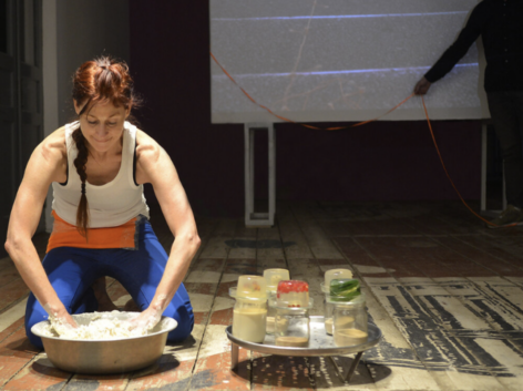 A photo of a light skinned Indigenous female presenting person faces forward and kneels in the left side of the photo. Their head is bent downward with their eyes almost closed. They have dark reddish hair pulled back in a long braid. The braid dangles forward over their rounded shoulders as they lean into a silver bowl. This is moira, their elbows are bent and their hands are in the bowl. The bowl is filled with white colored gooey dough that is drying in large chunks on their hands. They are wearing a white sleeveless tank top, a wide bright orange a pouch at their bent waist and bright blue pants. The bowl sits on a wooden plank floor that has large painted  and scuffed shapes in white, black and brown. On moira’s right is a silver tray with holes. It casts a round shadow with dots. On top of the tray are 5 clear glass jars partly filled with gooey liquids and powdery dry grains. The jars have clear gelatin in rounded shapes on top. Each gelatin visible has long green narrow leaves, bright red berries, or a tiny speckled brown egg inside. Behind moira is part of a video projection on a screen. The projection is a close up bird's eye view of a worn stone staircase with a bright orange string slowly moving up the stairs. In front of and to the right of the screen is a live person’s arm stretching in front of the screen and gently holding a bright orange ribbon in their brown hand. The ribbon is attached to the screen and it hangs in 2 big U’s as it dribbles onto the floor. The person appears as a silhouette dressed in black, visible from the shoulders down. Photo by Yuli Dudnyk. 