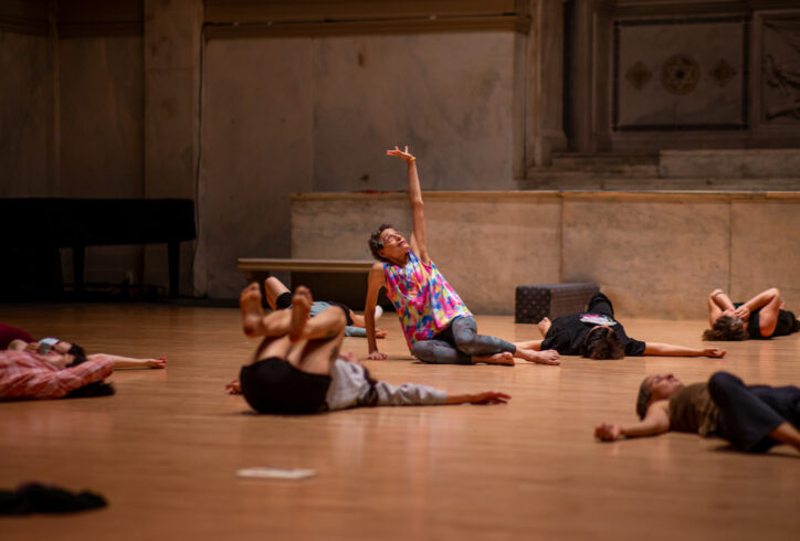 Graphic with text over an image. Barbara Mahler teaches a class. She sits on the floor with one arm reaching for the sky, her gaze following. Students surrounding her lie with their backs flat on the floor and knees bent. Photo by Rachel Keane.