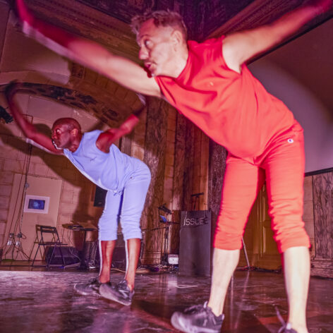 Neil Greenberg and Paul Hamilton in performance. On the left Paul wears all blue and on the right Neil wears all red. The balance on their heels sending their hips backward and their right arm forward to counter balance. Courtesy of ISSUE Project Room. Photography by Cameron Kelly McLeod