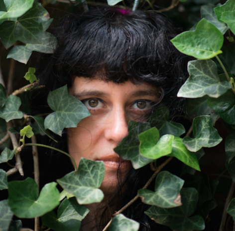 Nicole, a white person with dark, wavy hair and green eyes looks directly at the camera with a serious expression. The photograph is taken outdoors, up close, and her face is partially obscured by tendrils of ivy. Photo by Kenzie Crash