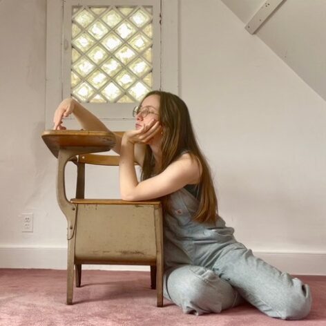 Chloe wears pinstriped overalls and glasses. She has white skin and long brown hair. She kneels on pink carpeted floor and leans onto an old fashioned school desk. One hand holds her chin and the other reaches lazily onto the desktop.