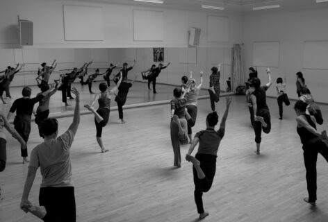 A large class with dancers seen from the back holding the tops of their right foot with their left hands while their right arms gesture to the high diagonal. Image by Tim Russell.