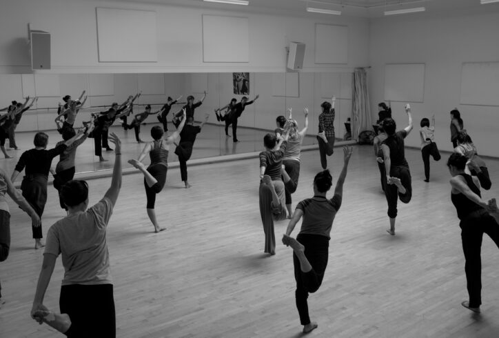 A large class with dancers seen from the back holding the tops of their right foot with their left hands while their right arms gesture to the high diagonal. Image by Tim Russell.