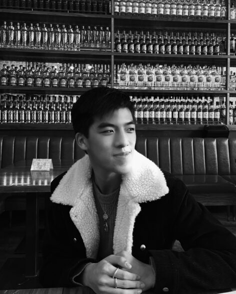 Black and white portrait of Ethan gazing and smiling off-camera. He is wearing a sherpa jacket, a grey sweater, two necklaces, and two rings. His hands are clasped.