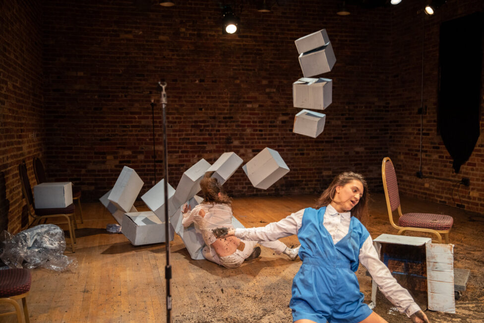 Surrounded by white cardboard boxes, convention-style chairs, and dirt, Lena Engelstein and Lisa Fagan perform. Photo credit to Walter Wlodarczyk.