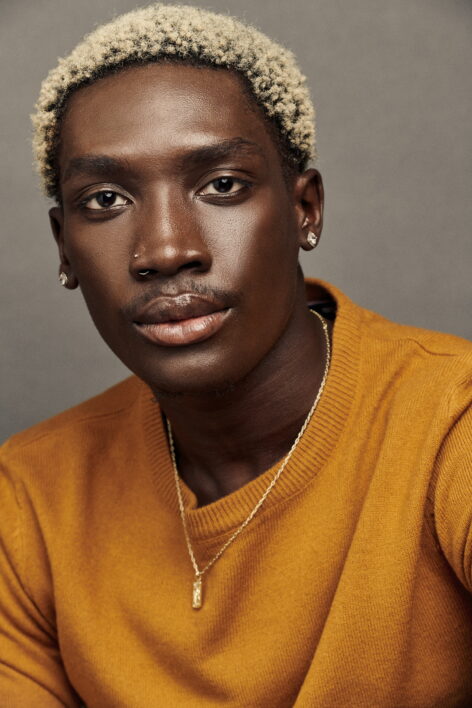 A photo of Babou Sanneh looking into the camera. He has blonde bleached short afro hair and wears a burnt orange top. Photo courtesy of the artist.