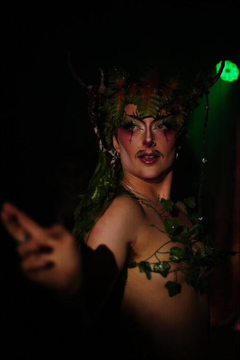 Lux as the Green Man wears leaves on his torso and in his long green wig. He gestures towards the camera, summoning viewers into the woods with him. Photo courtesy of the artist.