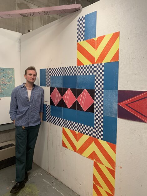 Blonde man wearing a blue shirt and green pants stands to the left of a vibrant installation featuring square, mosaiced prints of geometric patterns.