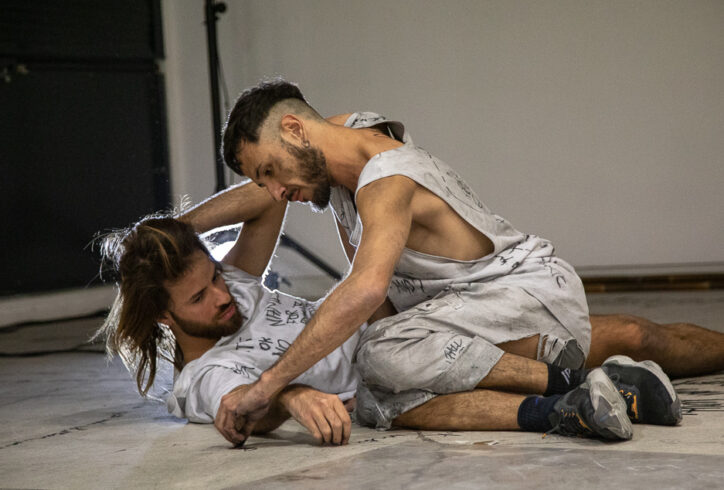 Two male dancers wearing sneakers and white practice clothes with writing and drawings in black charcoal grapple on a grey floor. Photo by Hans Peter Ibaghino.