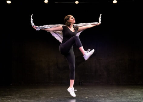 Leila Mire wears all black and hops, lifting one leg toward her chest. She is draped in a black and white keffiyeh and performs against a black backdrop. Photo by Steven Pisano.