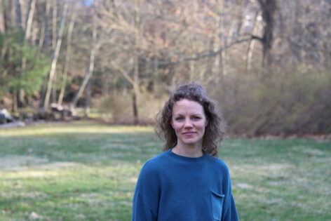 Marilyn Maywald Yahel, a white woman with brown curly hair, stands in front of grass and trees. She wears a blue sweater and looks into the camera with a slight smile. Photo by Sam Yahel