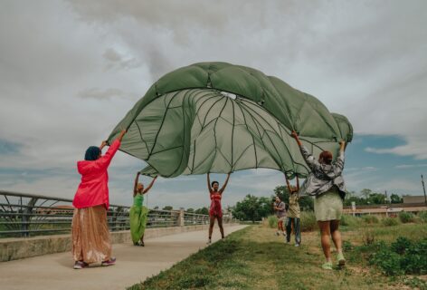 Photo of performers with a green parachute. They lift it into the air as it bellows. The sky is grey and they are dressed in bright colors. Image courtesy of the artist and DiverseWorks.