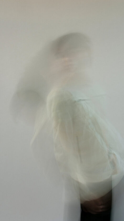 Long exposure portrait of Cameron in motion. They are falling against a white background. They have short brown hair and wear a white jacket with black pants. Photo by Mimi Kim.