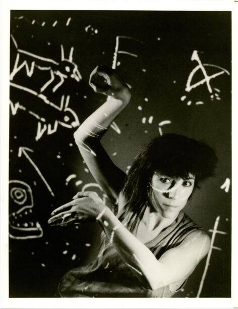 A black and white photo of Jo Andres. Jo reaches one arm up bending at the wrist and elbow. The other arm reaches across the chest also bending at the elbow. They look beyond the camera. The image has several line drawings and doodled of animal motifs and other miscellaneous markings. Photo by Paula Court.